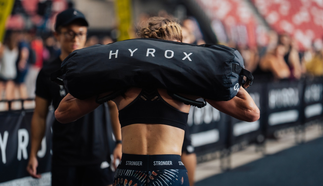 HYROX athlete carrying a weighted HYROX sandbag during a race workout station.