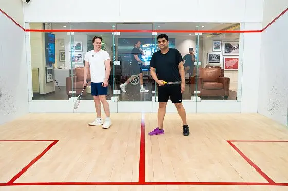 Two men playing squash in an indoor court with glass walls and red line markings on the wooden floor.