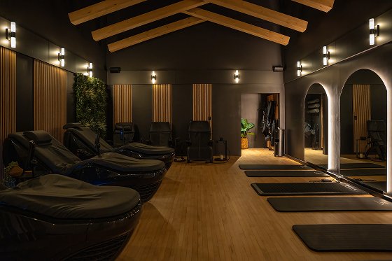 Modern spa and wellness studio with yoga mats, massage chairs, wooden beams, and ambient lighting.