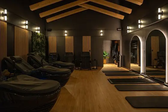 Modern spa and wellness studio with yoga mats, massage chairs, wooden beams, and ambient lighting.