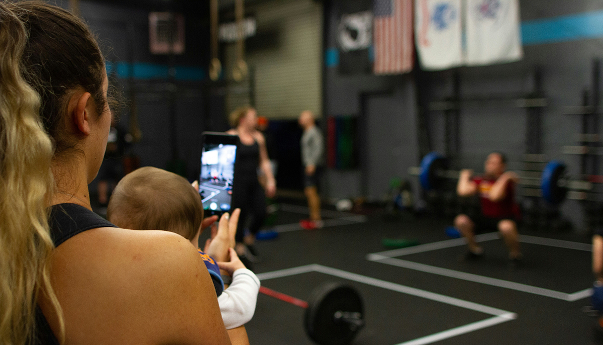 A person holding a baby while watching and recording a workout happening in a gym.