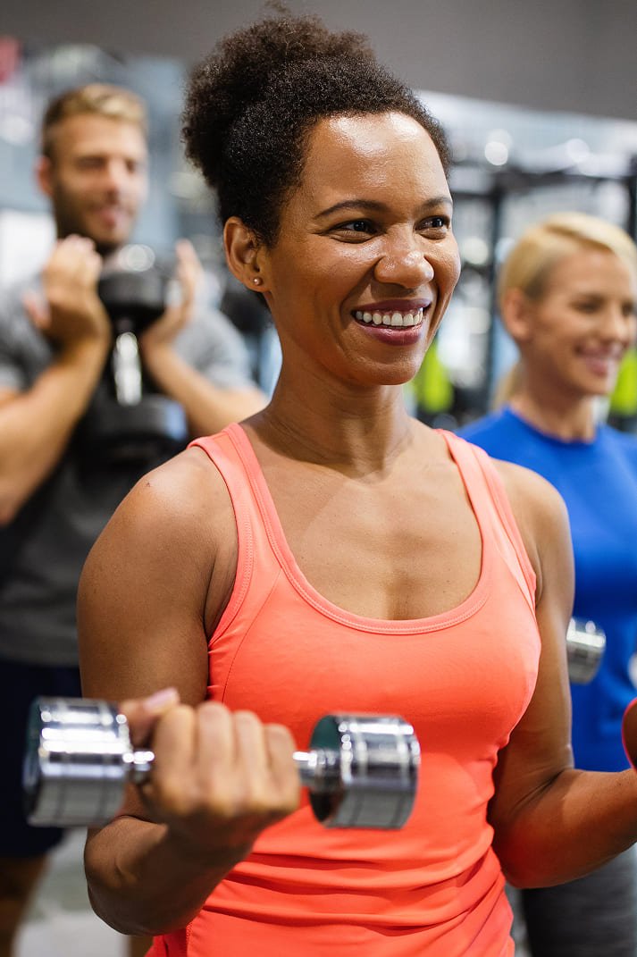 Woman in coral tank top smiling while holding dumbbells during a group fitness class at a gym.