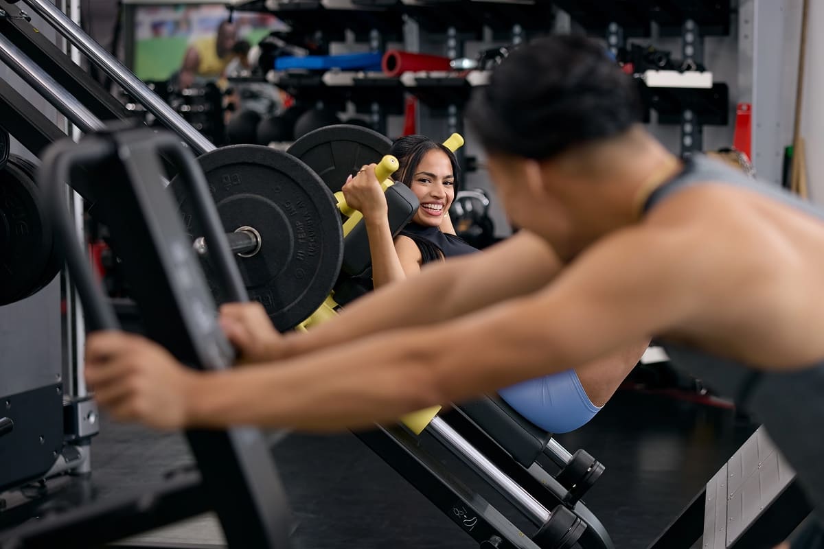 A woman smiling while using a leg press machine in a gym, with another person working out on a nearby machine in the foreground.