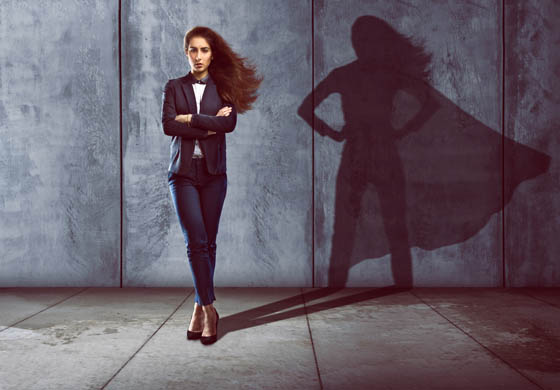 Professional young woman in a suit standing with arms and legs crossed, shadow on the wall behind is her but wearing a cape