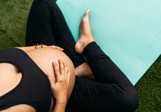 Close-up of pregnant woman's belly as she sits on the side of a yoga mat on the grass
