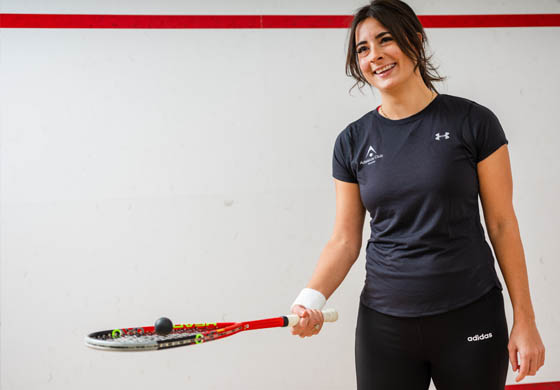 Young female squash player poses with her racquet out and a squash ball balanced on top on the squash courts at the Adelaide Club