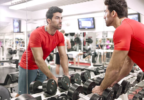 Young, attractive, fit man in red t-shirt looking at himself in the mirror as he grabs dumbbells at the Adelaide Club