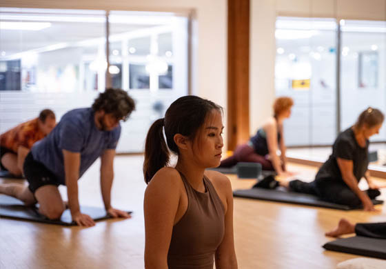 Young Asian woman in the forefront mid-yoga workout in the Flow Studio at the Adelaide Club. Other members and the instructor are featured stretching in the background.