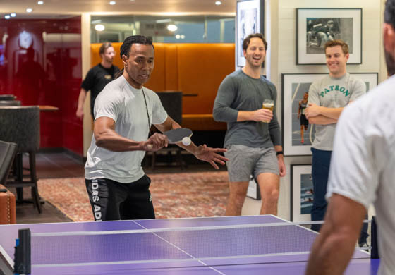 Middle aged black man playing table tennis in the WorkHUB at the Adelaide Club against another fit man. Two other men stand in the background leaning against the wall, drinking beers, and watching the ping pong action.