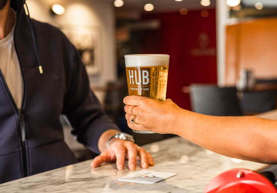 Arm of female bartender handing a pint glass of beer across the bar to a man in a blue zip-up hoodie